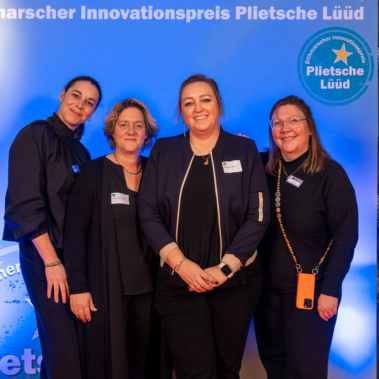 Vier Personen stehen vor einem blauen Hintergrund mit der Aufschrift "Dithmarscher Innovationspreis Plietsche Lüüd". Sie lächeln in die Kamera und tragen dunkle Kleidung.