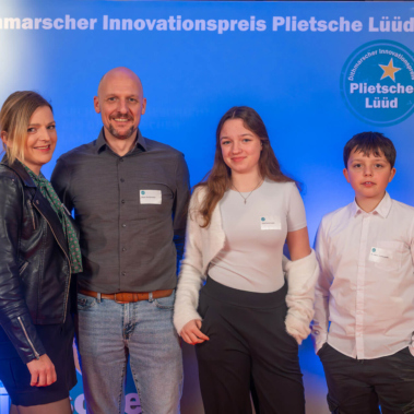 Vier Personen stehen vor einer blauen Wand mit der Aufschrift "Dithmarscher Innovationspreis Plietsche Lüüd". Sie tragen Namensschilder und lächeln in die Kamera. Auf einem Tisch rechts liegen Kissen übereinander.