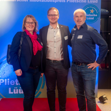 Eine Frau und zwei Männer stehen auf einem roten Teppich vor einer blauen Wand mit der Aufschrift "Dithmarscher Innovationspreis Plietsche Lüüd". Alle drei tragen Jeans und Blazer und lächeln in die Kamera.