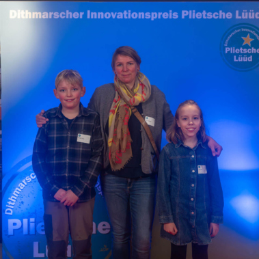 Eine Frau steht zwischen einem Jungen und einem Mädchen vor einem blauen Hintergrund mit dem Text "Dithmarscher Innovationspreis Plietsche Lüüd". Alle drei lächeln in die Kamera und tragen Namensschilder.