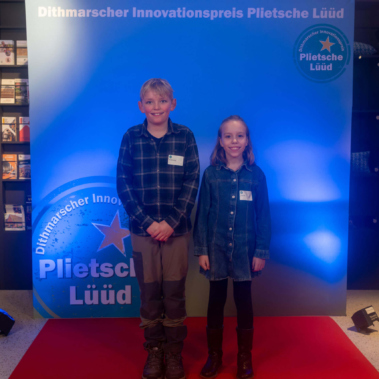Zwei Kinder stehen auf einem roten Teppich vor einem blauen Hintergrund mit dem Logo des Dithmarscher Innovationspreises „Plietsche Lüüd“. Beide tragen Namensschilder und lächeln in die Kamera.