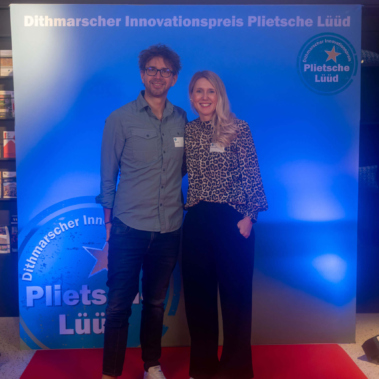 Ein Mann und eine Frau stehen auf einem roten Teppich vor einer blau beleuchteten Kulisse mit der Aufschrift "Dithmarscher Innovationspreis Plietsche Lüüd". Beide lächeln in die Kamera.