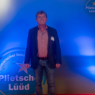 Ein Mann steht auf einem roten Teppich vor einem blau beleuchteten Hintergrund mit dem Text "Dithmarscher Innovationspreis Plietsche Lüüd". Er trägt ein Sakko, ein Hemd und Jeans.
