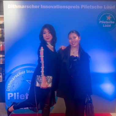 Zwei Personen stehen auf einem roten Teppich vor einer blauen Wand, die den Dithmarscher Innovationspreis Plietsche Lüüd zeigt. Beide lächeln; die linke Person hebt ein Bein an und hält eine Tasche, die rechte Person hat ihre Hände vor sich gefaltet.