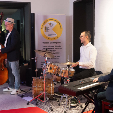 Drei Musiker spielen zusammen. Links steht ein Mann mit einem Kontrabass, in der Mitte sitzt ein Mann am Schlagzeug, und rechts spielt ein Mann Klavier. Sie sind in einem Raum mit weißer Wand und einem Schild.