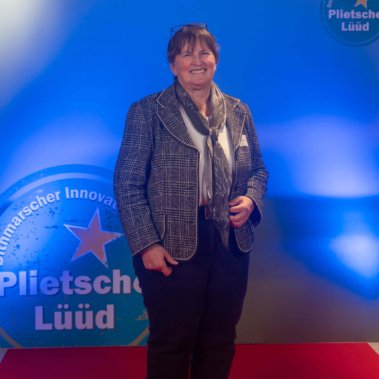 Eine Person steht auf einem roten Teppich vor einer blauen Wand mit dem Logo des Dithmarscher Innovationspreises "Plietsche Lüüd". Sie trägt eine karierte Jacke und einen Schal.