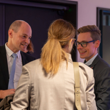 Drei Personen in Anzügen stehen in einem geschlossenen Raum und unterhalten sich freundlich. Eine Person ist von hinten zu sehen, die anderen stehen sich gegenüber. Die Stimmung wirkt professionell.