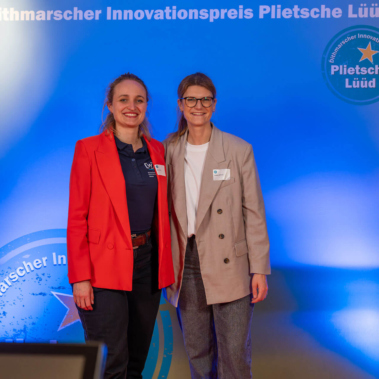 Zwei Personen stehen vor einem blauen Hintergrund mit der Aufschrift "Dithmarscher Innovationspreis Plietsche Lüüd". Beide lächeln in die Kamera. Links trägt rot, rechts trägt beige. Im Hintergrund sind Regale sichtbar.