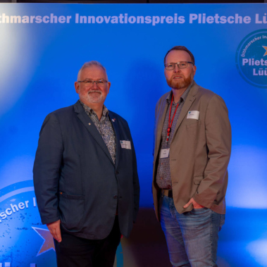 Zwei Männer stehen vor einer blauen Wand mit der Aufschrift "Dithmarscher Innovationspreis Plietsche Lüüd". Beide tragen Sakkos und Hemden. Verschiedene Broschüren sind im Hintergrund zu sehen.