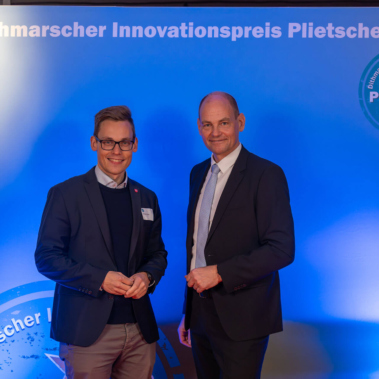 Zwei Männer in Anzügen stehen vor einem blau beleuchteten Hintergrund mit der Aufschrift "Dithmarscher Innovationspreis Plietsche Lüüd". Beide lächeln in die Kamera. Im Hintergrund sind Bücherregale erkennbar.