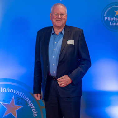 Eine lächelnde Person steht vor einem blauen Hintergrund mit der Aufschrift „Deutscher Innovationspreis Plietsche Lüüd“. Die Person trägt einen dunklen Anzug und ein hellblaues Hemd.