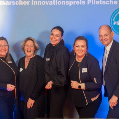 Eine Gruppe von sechs Personen steht lächelnd vor einem blauen Hintergrund mit der Aufschrift "Dithmarscher Innovationspreis Plietsche Lüüd." Die Personen tragen legere bis formelle Kleidung.