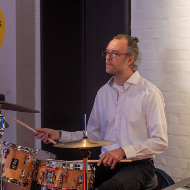 Ein Mann in weißem Hemd sitzt an einem Schlagzeug und spielt. Er blickt konzentriert nach vorne. Die Wand hinter ihm ist weiß, und links ist ein Plakat teilweise sichtbar.
