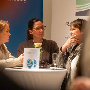 Drei Personen sitzen an einem runden Stehtisch und unterhalten sich. Auf dem Tisch stehen ein Blumengesteck und ein Glas. Im Hintergrund sind unscharfe Banner und eine Person am Rand sichtbar. Die Stimmung wirkt konzentriert.