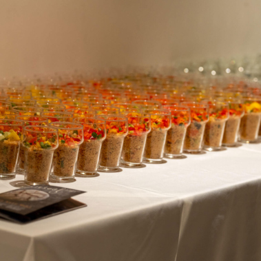 Auf einem weißen Tisch stehen viele kleine Gläser in Reihen. Die Gläser sind mit einer bunten Mischung aus Quinoa, Gemüse und Kräutern gefüllt. Im Vordergrund liegen zwei Karten. Der Hintergrund ist unscharf.