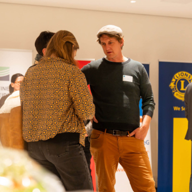 Gruppe von Menschen bei einer Veranstaltung in einem Konferenzraum. Zwei unterhalten sich lebhaft, andere stehen an Stehtischen. Im Hintergrund sind Banner verschiedener Organisationen zu sehen. Stimmung ist geschäftig und freundlich.