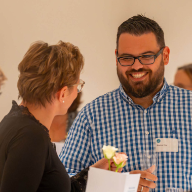 Ein Mann und eine Frau stehen in einem hellen Raum und unterhalten sich lächelnd. Der Mann trägt ein kariertes Hemd, die Frau ein schwarzes Oberteil. Im Hintergrund sind weitere Personen unscharf zu sehen.