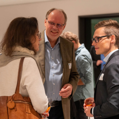 Eine Gruppe von Menschen steht in einem Raum und unterhält sich. Ein Mann trägt einen Anzug, während ein anderer Mann ein Jackett trägt. Eine Frau mit einer Tasche steht ihnen gegenüber. Andere Personen sind im Hintergrund sichtbar.