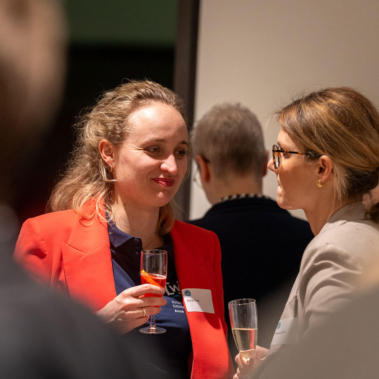 Zwei Frauen stehen in einem Innenraum und unterhalten sich. Eine Frau trägt einen roten Blazer, die andere eine beige Jacke. Beide halten Gläser in der Hand. Weitere Personen sind verschwommen im Hintergrund.