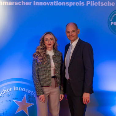 Zwei Personen stehen vor einem blauen Hintergrund mit der Aufschrift „Dithmarscher Innovationspreis Plietsche Lüüd“. Links eine Frau mit langem Haar im Blazer, rechts ein Mann im Anzug. Beide lächeln in die Kamera.
