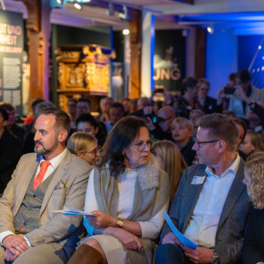 Eine Gruppe von Menschen sitzt eng zusammen in einem beleuchteten Raum. Sie tragen überwiegend formelle Kleidung und halten Programme. Im Hintergrund sind unscharfe Schautafeln und Dekorationen zu sehen.