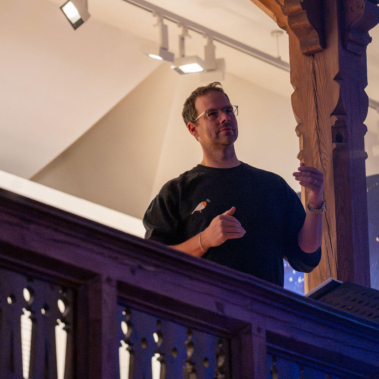 Ein Mann steht auf einem Balkon und spricht gestikulierend. Er trägt eine Brille und ein dunkles Oberteil. Der Hintergrund ist beleuchtet, mit Bildern und einer Holzkonstruktion an der Seite.