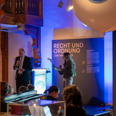 Ein Mann und eine Frau stehen auf einer Bühne in einem Raum mit dunkler Holzeinrichtung. Hinter ihnen ist eine blaue Wand mit der Aufschrift "Recht und Ordnung". Im Vordergrund sind Ausstellungsstücke in Glasvitrinen zu sehen.