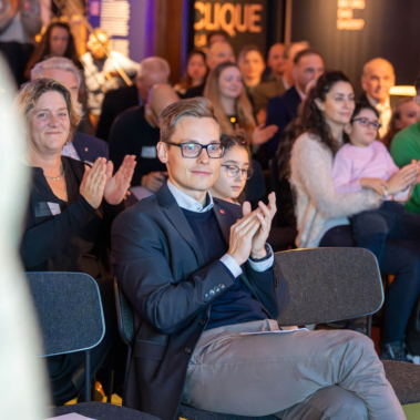 Personen sitzen auf Stühlen in einem Raum und applaudieren. Im Vordergrund klatscht eine Person mit Brille, dahinter sind weitere Personen zu sehen. Der Raum ist gut gefüllt und beleuchtet.