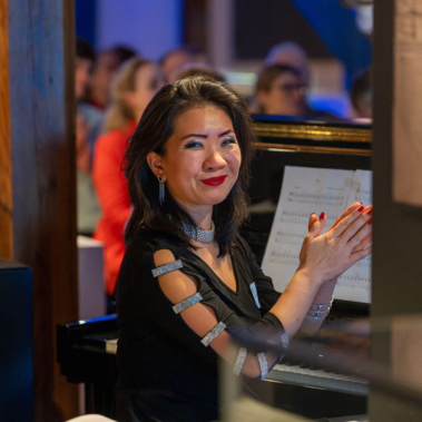 Eine Frau sitzt an einem Klavier und klatscht. Sie trägt ein schwarzes Kleid mit glitzernden Details. Der Hintergrund zeigt unscharfe Menschen in einem beleuchteten Raum.