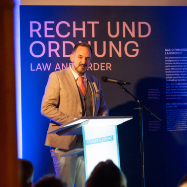 Ein Mann steht an einem Rednerpult und spricht in ein Mikrofon. Hinter ihm ist ein blau beleuchtetes Plakat mit der Aufschrift "RECHT UND ORDNUNG" sichtbar. Der Raum ist warm beleuchtet.