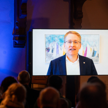 Ein Mann in Anzug und Brille spricht auf einem Bildschirm vor einem gemischten Publikum in einem gedämpft beleuchteten Raum. Hinter ihm ist ein Bild mit bunten Segeln zu sehen.