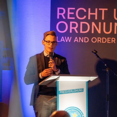 Ein Mann steht an einem Rednerpult und spricht in ein Mikrofon. Hinter ihm sind die Wörter "Recht und Ordnung" auf einer Wand zu sehen. Der Raum ist beleuchtet und es findet eine Veranstaltung statt.