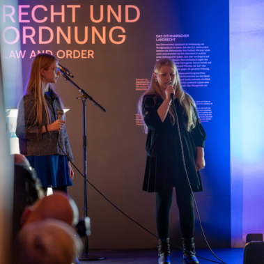 Zwei Personen sprechen in Mikrofone auf einer Bühne, umgeben von blau-violettem Licht. Im Hintergrund ist Text auf einer Wand mit der Aufschrift „Recht und Ordnung“ zu sehen. Zuschauer sitzen im Vordergrund.