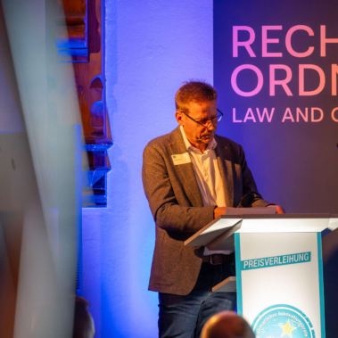 Ein Mann steht an einem Rednerpult und spricht. Im Hintergrund ist ein Schild mit der Aufschrift "RECHT UND ORDNUNG" und "LAW AND ORDER" zu sehen. Er trägt einen Anzug mit offenem Hemd. Beleuchtung ist warm.