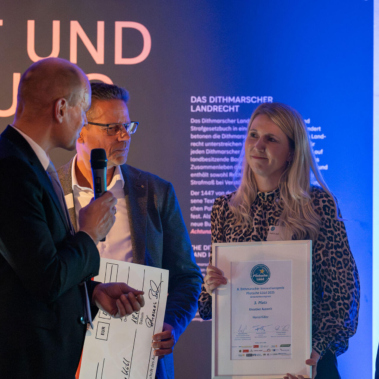 Vier Personen stehen vor einer beleuchteten Wand. Eine Frau hält eine Urkunde und ein Mann überreicht einen großen symbolischen Scheck. Mikrofone und ein Rednerpult sind ebenfalls im Bild zu sehen.