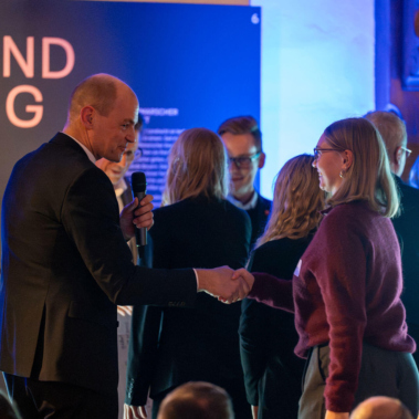 Eine Gruppe von Menschen steht in einem beleuchteten Raum. Eine Person im Anzug schüttelt einer anderen die Hand. Im Hintergrund sind weitere Personen und ein Rednerpult mit der Aufschrift "Preisverleihung".