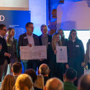 Eine Gruppe von Männern und Frauen steht auf einer Bühne. Eine Person hält einen symbolischen Scheck, eine andere ein Zertifikat. Im Hintergrund sind Banner und eine Projektion zu sehen. Zuschauer sitzen im Vordergrund.