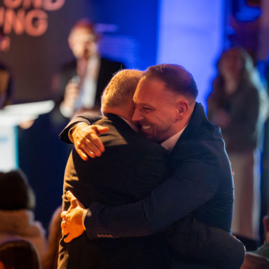 Zwei Männer umarmen sich herzlich in einem Raum mit mehreren Personen. Im Hintergrund sind unscharfe, beleuchtete Schilder und Menschen zu erkennen. Die Szene wirkt warm und freundschaftlich.