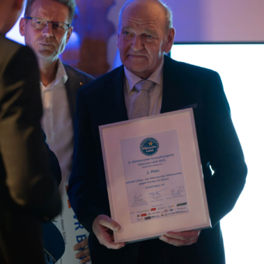 Eine Person im Anzug hält eine gerahmte Urkunde. Im Hintergrund steht eine weitere Person. Die Szene ist in ein sanftes bläuliches Licht getaucht.