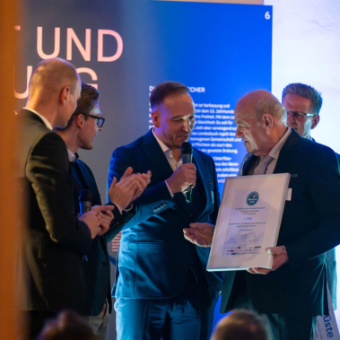 Eine Gruppe von Männern in Anzügen steht bei einer Veranstaltung. Ein Mann hält eine gerahmte Urkunde, während andere applaudieren. Die Szene ist in warmem, blauem Licht beleuchtet.