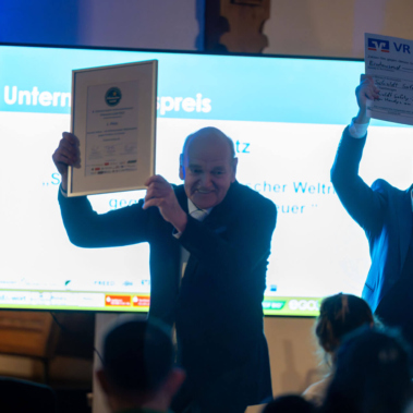 Eine lächelnde Person hält eine gerahmte Urkunde hoch. Eine andere Person daneben hält einen großen Scheck. Im Hintergrund sind ein Bildschirm und Zuschauer erkennbar. Die Szene wirkt festlich und freudig.