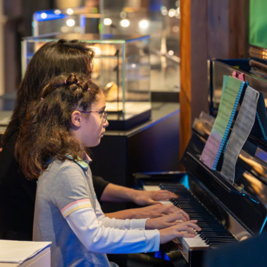 Ein Kind und eine erwachsene Person sitzen nebeneinander an einem Klavier und spielen. Beide sind von hinten zu sehen. Im Hintergrund befinden sich Vitrinen und eine leuchtende Anzeige.