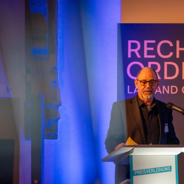 Ein Mann steht an einem Podium und spricht. Hinter ihm steht ein Schild mit der Aufschrift "Recht und Ordnung". Der Raum ist in blauen und orangen Tönen beleuchtet.