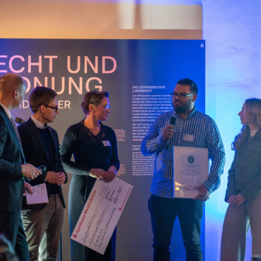 Eine Gruppe von Personen steht auf einer Bühne vor einem Schild mit der Aufschrift "Recht und Ordnung". Eine Person spricht in ein Mikrofon. Andere halten Schecks und Urkunden. Die Szene ist in blaues Licht getaucht.