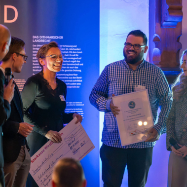 Eine Gruppe von Menschen steht in einem beleuchteten Raum. Zwei Personen halten große Schecks und Dokumente. Eine Person spricht in ein Mikrofon. Alle lächeln und tragen formelle Kleidung. Im Hintergrund ist eine blaue Wand mit Text.