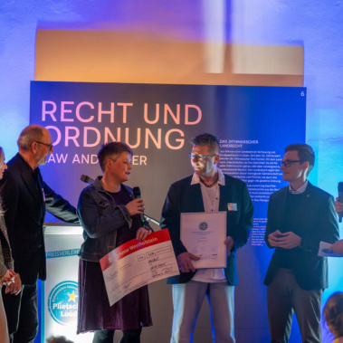 Fünf Personen stehen auf einer Bühne und halten einen großen symbolischen Scheck und Zertifikate. Im Hintergrund ist ein Schild mit der Aufschrift "Recht und Ordnung" sichtbar. Die Szene ist beleuchtet.