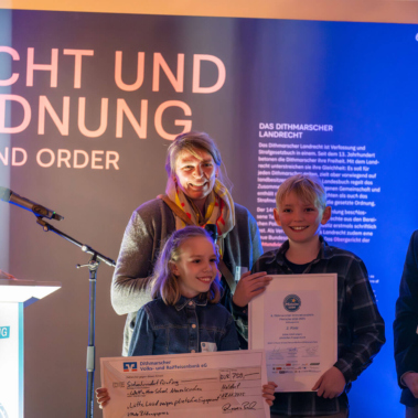 Eine Gruppe von Menschen steht vor einer blau beleuchteten Wand mit der Aufschrift "Recht und Ordnung". Zwei Personen in der Mitte halten einen großen Scheck und eine Urkunde, während andere Personen daneben stehen.