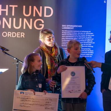 Eine Frau und drei Männer stehen auf einer Bühne mit einem Mikrofon. Zwei Personen halten Urkunden und einen großen Scheck. Im Hintergrund ist eine leuchtende Tafel mit Text. Die Szene wirkt wie eine Preisverleihung.