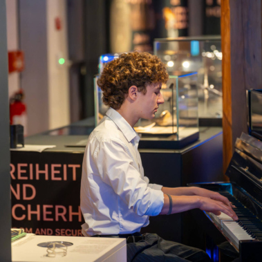 Ein Junge mit lockigem Haar spielt in einem Museum Klavier. Er trägt ein weißes Hemd. Im Hintergrund sind Ausstellungsvitrinen und ein Schild mit Text zu sehen. Die Umgebung wirkt modern und gut beleuchtet.