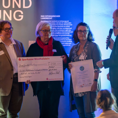 Vier Personen stehen auf einer Bühne, drei davon halten einen großen Scheck und eine Urkunde. Eine Person rechts spricht in ein Mikrofon. Im Hintergrund sind ein Logo und Text auf einer beleuchteten Wand zu sehen.